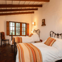 Experience Cowgirl Yoga™ - Argentina in a cozy bedroom with two single beds, striped blankets and pillows, wooden beams, bookshelf, wall lamps, and sunlight flooding through a window overlooking trees.